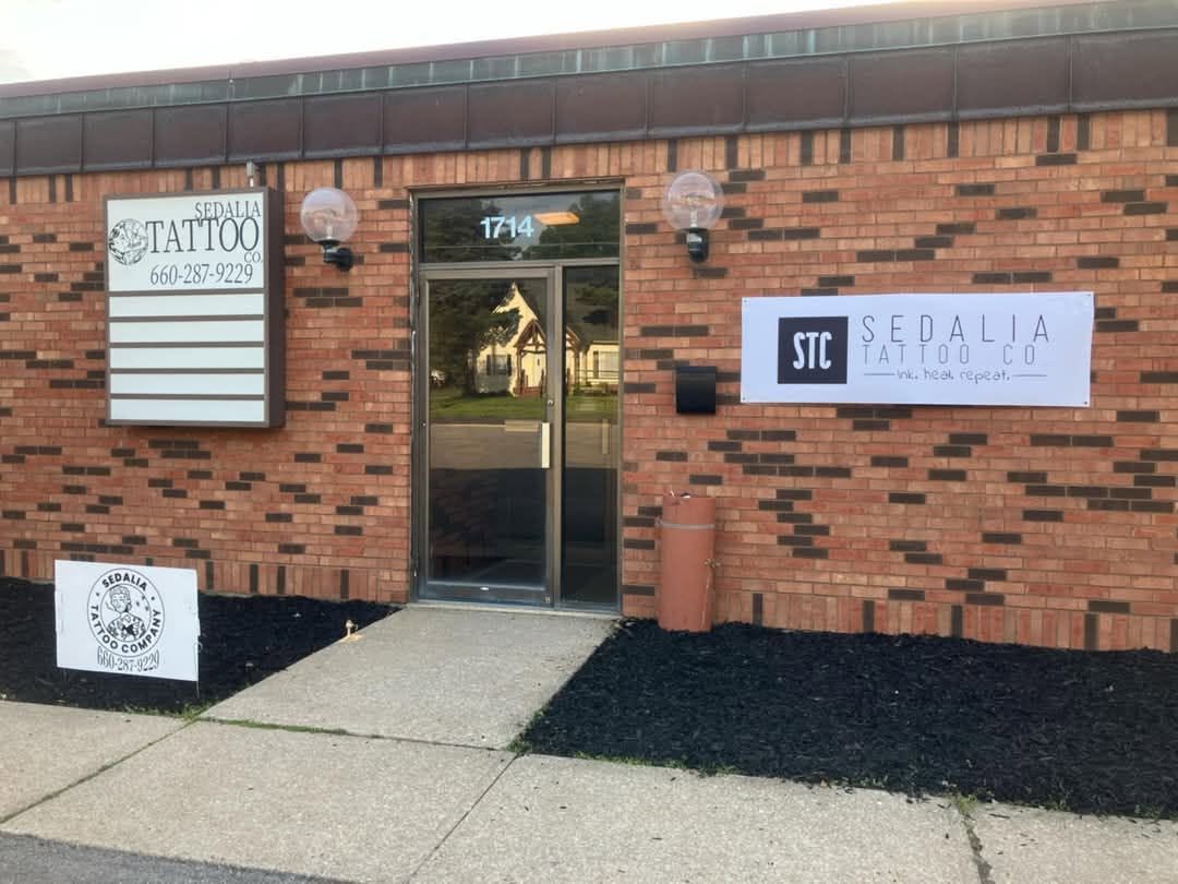 The Sedalia Tattoo Company shopfront: a brick unit with a glass door numbered 1714, a lit sign box reading Sedalia Tattoo Co with the phone number, and a banner reading ink. heal. repeat.