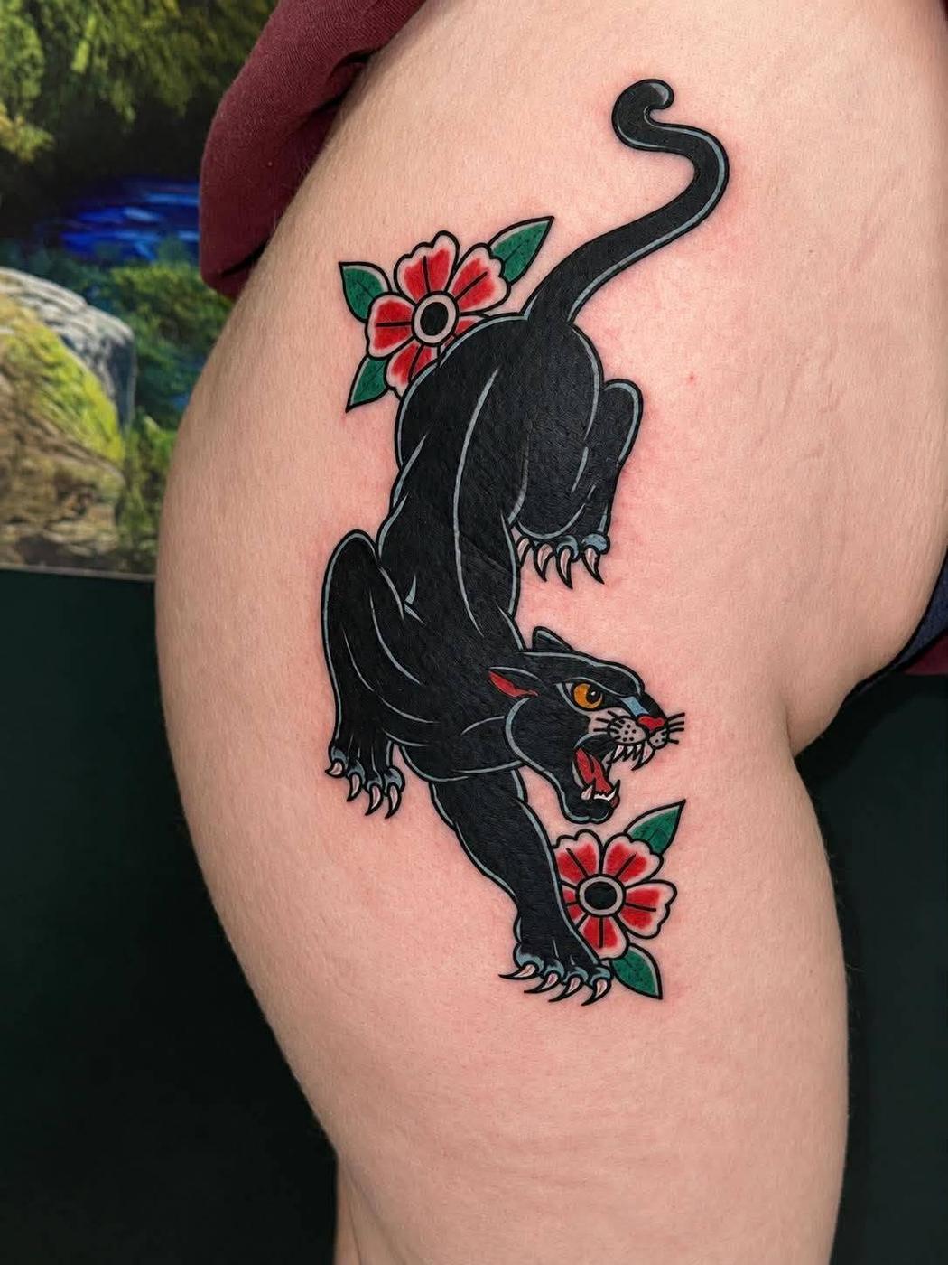 A crouching black panther with red and green flowers, tattooed in full color in a traditional style on a thigh.